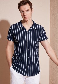 Maglietta a righe blu navy e bianca a maniche corte con colletto a punta e chiusura a bottoni, realizzata in un tessuto leggero e testurizzato.