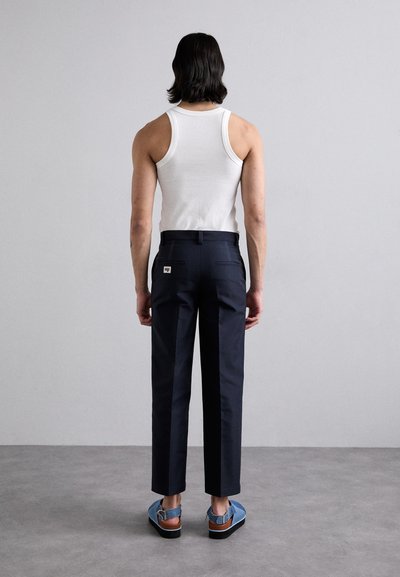 Navy tailored pants with a slim fit, paired with a white ribbed tank top and blue sandals with a brown strap. Plain background.
