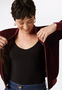 Giacca bomber bordeaux con zip e texture liscia in velluto, indossata su una canotta nera. Tasche laterali visibili, jeans in denim con bottone.