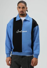 Sweat-shirt en mélange de coton bleu et noir, doté d'un col zippé, d'une coupe ample et du texte blanc « Saur essence » en écriture cursive sur le devant.