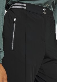 Svarta byxor i slät tyg har en dragkedjeficka, dekorativa knappar och ett midjeband med silverrandig detaljer.