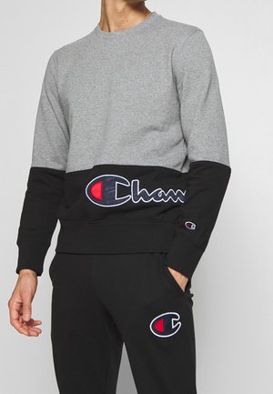 Mężczyzna noszący szarą i czarną bluzę Champion z logo na klatce piersiowej oraz czarne spodnie dresowe z logo na lewej udzie, ręka w kieszeni.