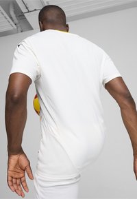 Camiseta atlética blanca de manga corta, con un acento amarillo en el cuello y un tejido texturizado. Una pelota roja y amarilla es parcialmente visible.