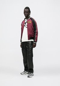 Mees seisab, kandes weinpunast ja musta pommitusjopet, valget graafilise mustriga dressipluusi, musti tööpükse ja valgeid-musta värvusega tossusid.