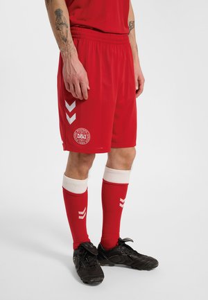 Mandlig atlet iført røde fodboldshorts og sokker med hvide chevrons og sorte fodboldstøvler, stående mod en ensfarvet baggrund.
