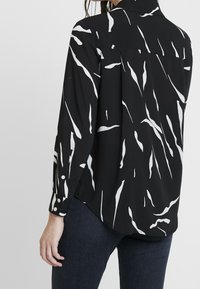 Schwarze Bluse mit weißen abstrakten Mustern, langen Ärmeln mit geknöpften Manschetten und geschwungener Saumlinie. Hergestellt aus leichtem, fließendem Stoff.