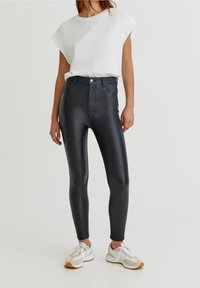 Leggings de imitación cuero negros de cintura alta con una textura suave, corte ajustado y un cierre frontal con botón visible, combinados con una camiseta blanca y zapatillas.