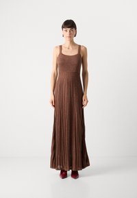 Maxikleid aus metallisch braunem Stoff, mit breiten Trägern, einem taillierten Oberteil und einem gefalteten, schwingenden Rock. Akzentuiert mit glänzend rotem Schuhwerk.