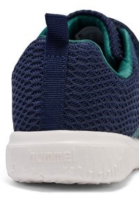 Marinblå mesh-idrottssko med gröna detaljer, med en strukturerad yta och en vit gummisula. Har en tygöglan vid hälen.