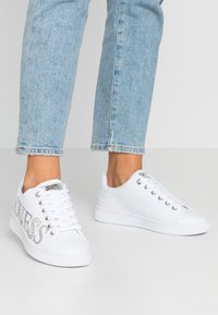 Baskets blanches en matériau synthétique lisse, avec des accents et des œillets argentés, ainsi que des lacets. Le design comprend des lettres décoratives.