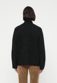 Personne aux cheveux courts et foncés portant un pull noir à manches longues en matière duveteuse et un pantalon en velours côtelé marron, vue de dos sur un fond blanc uni.