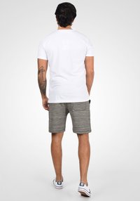 T-shirt blanc à manches courtes, shorts gris texturé et baskets blanches avec des accents bleus ; vu de dos, montrant un tatouage sur le bras.