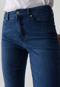 Jean en denim bleu foncé avec un design ajusté, doté d'une fermeture à boutons, d'accents en métal et de détails de couture subtils sur les poches.