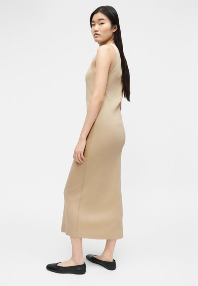 Γυναίκα με μακριά μαύρα μαλλιά που φοράει μπεζ midi αμάνικο φόρεμα και μαύρα φλατ παπούτσια, στέκεται μπροστά σε απλό λευκό φόντο.