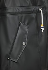 Black waterproof fabric with a diagonal pocket design, featuring a silver button and a drawstring with a colored accent. Textured surface.