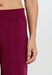 Joggings bordeaux avec une taille côtelée, présentant une texture lisse et une poche latérale. De subtils accents pailletés sont visibles.