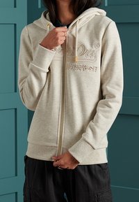 Felpa beige con zip e cappuccio regolabile, logo ricamato, polsini e orlo a costine, indossata sopra una maglietta scura su uno sfondo turchese.