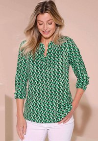 Blusa verde a fantasia geometrica, con abbottonatura frontale a metà e maniche tre quarti. Indossata con pantaloni bianchi.