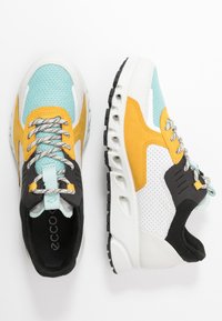 Sportskor med en nät- och mockadesign i turkos, gul, svart och vit. Har en vadderad krage och en strukturerad sula för bra grepp.