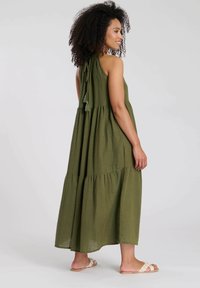 Olijfgroen, gelaagde maxi-jurk met hoge hals, gestrikte detail en textiel met structuur. Beschikt over franje en een vloeiende stijl.