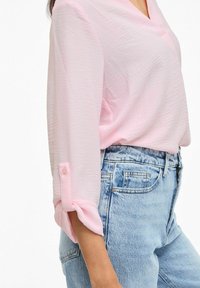 Femme portant un chemisier rose clair à manches longues retroussées et un jean denim taille haute bleu clair, debout devant un fond blanc.