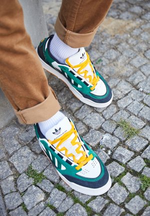 Osoba nosząca białe, zielone i granatowe buty sportowe Adidas z żółtymi sznurówkami i białymi skarpetkami stojąca na szarej brukowanej nawierzchni.