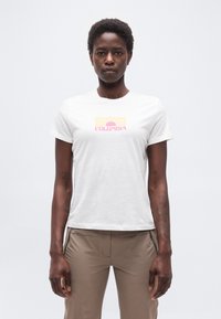 Γυναίκα που φοράει λευκό t-shirt Columbia με ροζ και κίτρινο γραφικό ήλιου, συνδυασμένο με ανοιχτό καφέ παντελόνι με φερμουάρ, στέκεται μπροστά σε απλό φόντο.