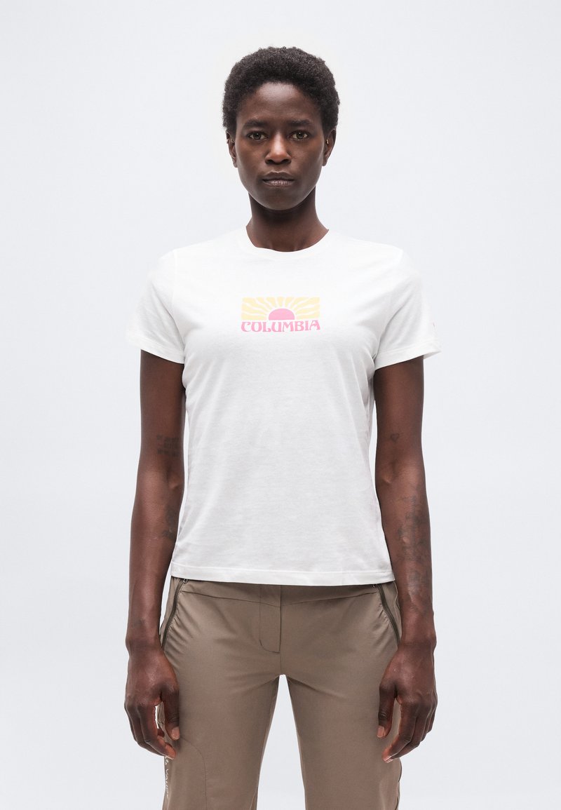 Γυναίκα που φοράει λευκό t-shirt Columbia με ροζ και κίτρινο γραφικό ήλιου, συνδυασμένο με ανοιχτό καφέ παντελόνι με φερμουάρ, στέκεται μπροστά σε απλό φόντο.