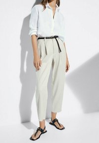 Camicia bianca in lino, pantaloni beige a vita alta con pieghe, cintura nera e sandali neri flat dal design minimale con dettagli dorati.