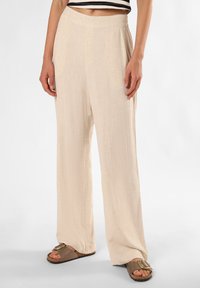 Beige Leinen-Hosen mit weitem Bein und glatter Textur, ausgestattet mit einem hohen Taillendesign. Kombiniert mit braunen Sandalen mit einem metallenen Schnallendetail.