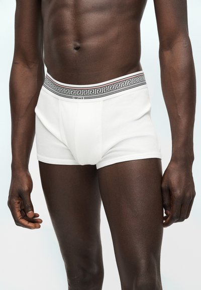 Modèle masculin portant un boxer blanc Versace avec une ceinture à motif clé grecque noire, debout devant un fond clair uni.