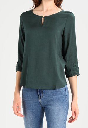 Femme portant un chemisier vert foncé à manches longues avec un petit bouton en forme de trou de serrure à l'avant et un jean bleu sur fond blanc.