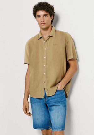 Άνδρας με σγουρά μαλλιά που φοράει μπεζ κοντομάνικο πουκάμισο με κουμπιά και μπλε τζιν σορτς, στέκεται μπροστά σε απλό φόντο.