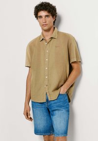 Άνδρας με σγουρά μαλλιά που φοράει μπεζ κοντομάνικο πουκάμισο με κουμπιά και μπλε τζιν σορτς, στέκεται μπροστά σε απλό φόντο.