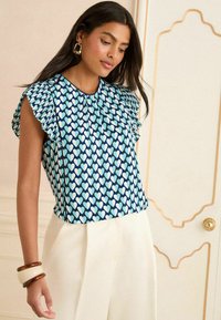 Blå kortärmad topp med ett hjärtmönster i ljusblått och vitt. Volangärmar och samlad halsringning. Kombinerad med ljusa byxor.
