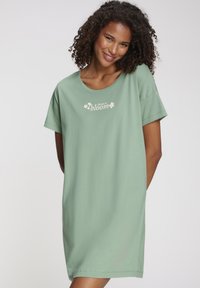 Žalia trumpomis rankovėmis suknelė iš minkštos medžiagos, su laisvu kirpimu. Turi apvalų kaklo iškirpimą ir priekyje esančią gėlių užrašą „choose to bloom“.