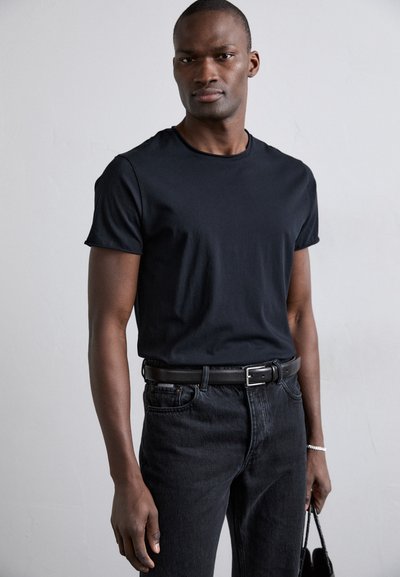 Camiseta de algodón negra de cuello redondo, de mangas cortas, combinada con jeans oscuros y un cinturón negro. La modelo sostiene un pequeño bolso de mano negro.