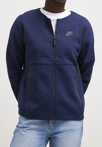 Mörkblå zip-up sweatshirt i mjukt tyg, med en liten Nike-logotyp på vänster bröst och två sidofickor med svarta detaljer.