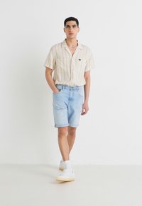 Cămașă scurtă cu mânecă scurtă, în dungi, într-o nuanță de bej și alb, asociată cu pantaloni scurți din denim bleu deschis. Modelul poartă adidași albi, stând în fața unui fundal simplu.