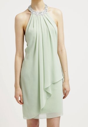 Mouwloze lichtgroene halterjurk met zilverkleurige kralenketting en gelaagde asymmetrische drapering aan de voorkant, gedragen door een staande persoon.