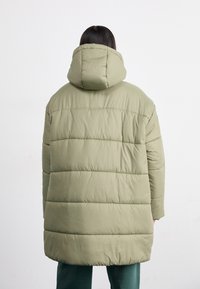 Chaqueta acolchada color verde oliva con capucha, que presenta un diseño acolchado y costuras horizontales. El material parece suave y aislante.