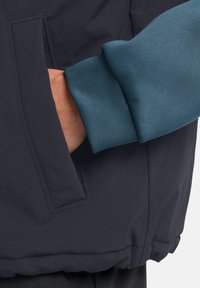 Marineblaue Jacke mit einem türkisfarbenen Manschetten. Verfügt über eine Seitentasche mit Klappe und einem glatten, wasserabweisenden Material. Gestickte Details sind sichtbar.