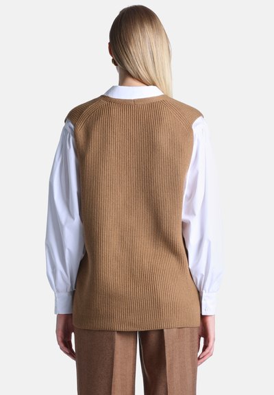 Femme vue de dos portant un gilet tricoté marron sur une chemise blanche à manches longues et un pantalon marron assorti sur un fond uni.