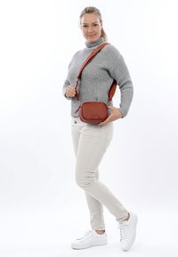 Naine hallis kampsunis, beežides pükstes ja valgetes tossudes, hoides pruunist nahast rihmpagasit valge tausta ees.