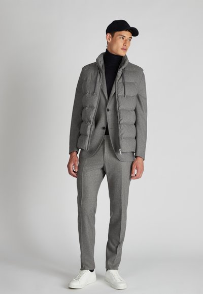 Grau gepolsterte Jacke über einem grauen Anzug, kombiniert mit einem schwarzen Rollkragenpullover und weißen Sneakern. Die Jacke hat einen hohen Kragen und Reißverschlussdetails.