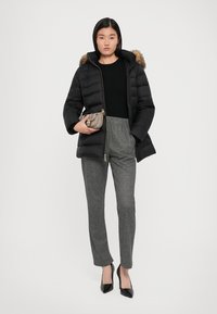 Zwarte pufferjack met een bontgevoerd capuchon, gecombineerd met een fitted zwart top en grijze geruite broek. Model houdt een kleine zilveren handtas vast.