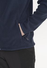 Marinblå fleecejacka med en dragkedjeficka och strukturerad tyg, bärs över svarta byxor, vilket visar en avslappnad design och praktiska detaljer.