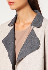 Lys beige uldfrakke med en mørkegrå krave. Designet har en blød tekstur og en åben front, lagd ovenpå en tan top.