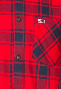 Chemise à carreaux rouge et noir avec une poche poitrine arborant un petit logo rectangulaire et deux boutons noirs visibles.