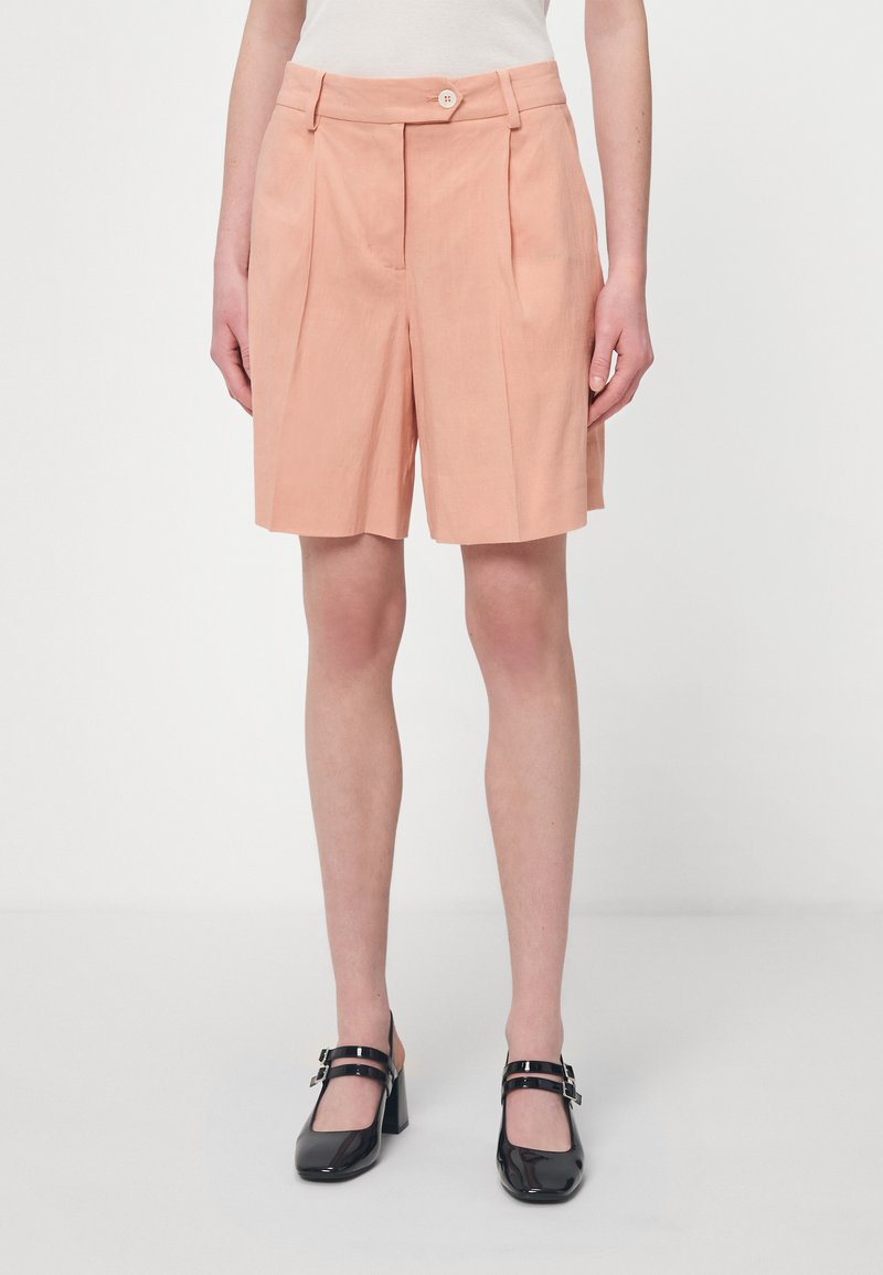 Pfirsichfarbene, plissierte Shorts mit einem Knopfverschluss. Der Stoff hat eine leinenartige Textur. Getragen mit schwarzen, patentreffenden Blockabsatzschuhen.